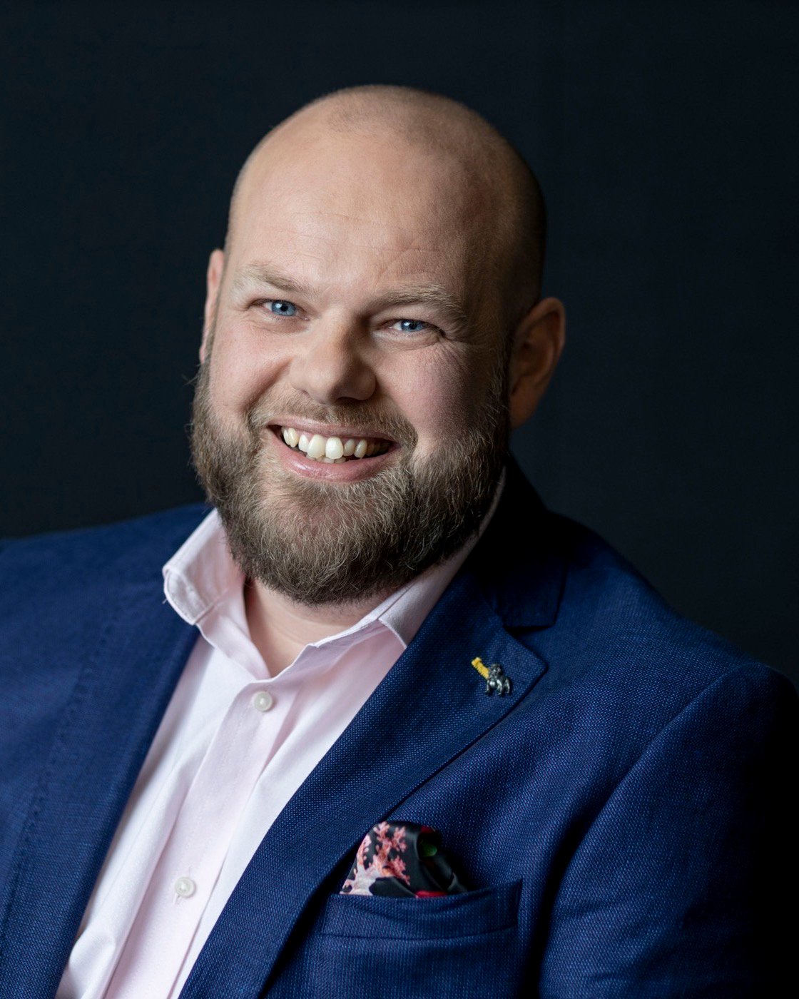 Ben Westlake, smiling, in a navy jacket against a dark studio background